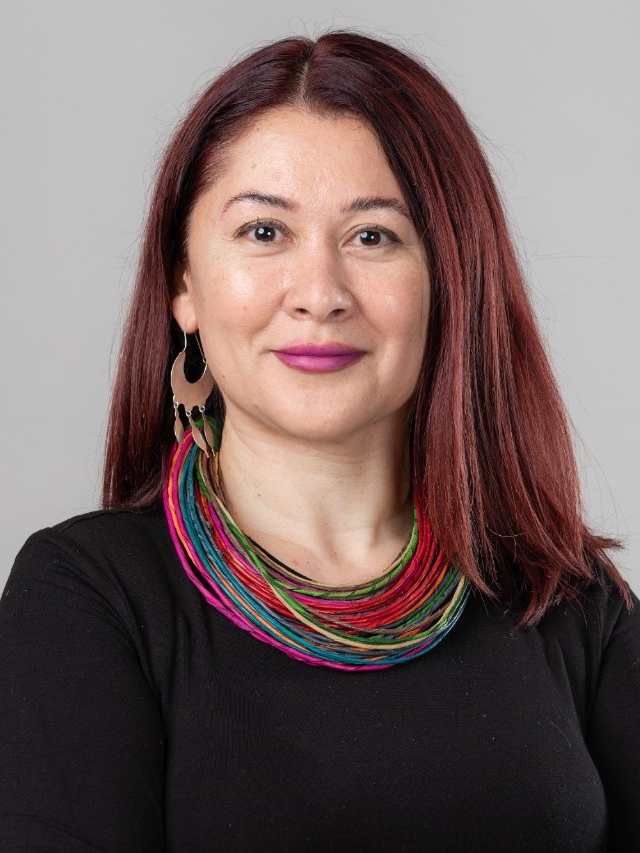 Dr. Evelyn Encalada Grez is wearing large silver earrings and a multicoloured necklace. She has dark red hair and is smiling at the camera.