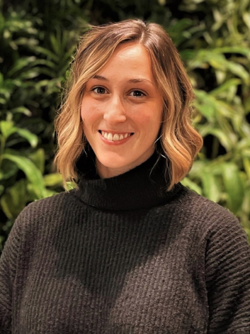 Rachelle is photographed in front of green plants that are blurred in the background. She has short wavy blonde hair, dark eyes, and is wearing a black sweater.