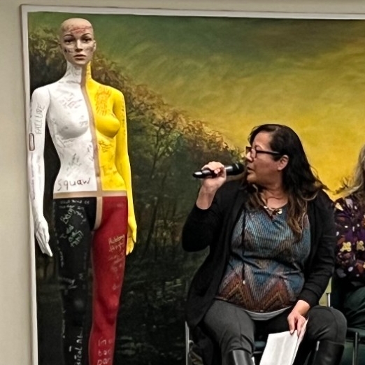 At the Shadbolt Fellow launch event in January 2023, Wanda sits on the stage and talks about her project referencing the mannequin next to her. The mannequin is coloured like a medicine wheel and has words and phrases written all over its body.