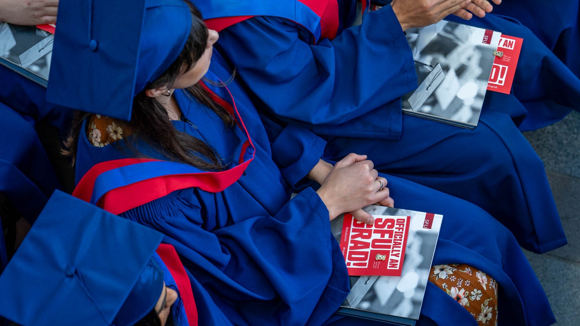 A graduate wearing blue and red regalia is photographed from above. They are holding a placard with a pin attached that reads OFFICIALLY AN SFU GRAD!