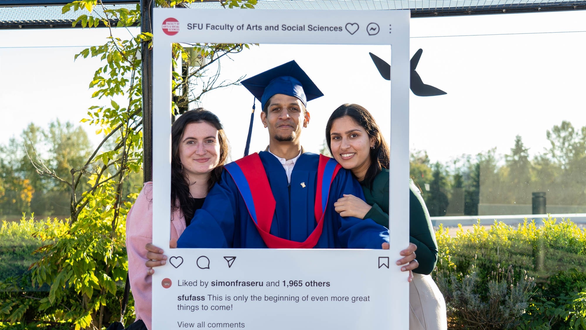 A FASS graduate in blue and red regalia poses outside with an SFU FASS Instagram frame with their two guests.