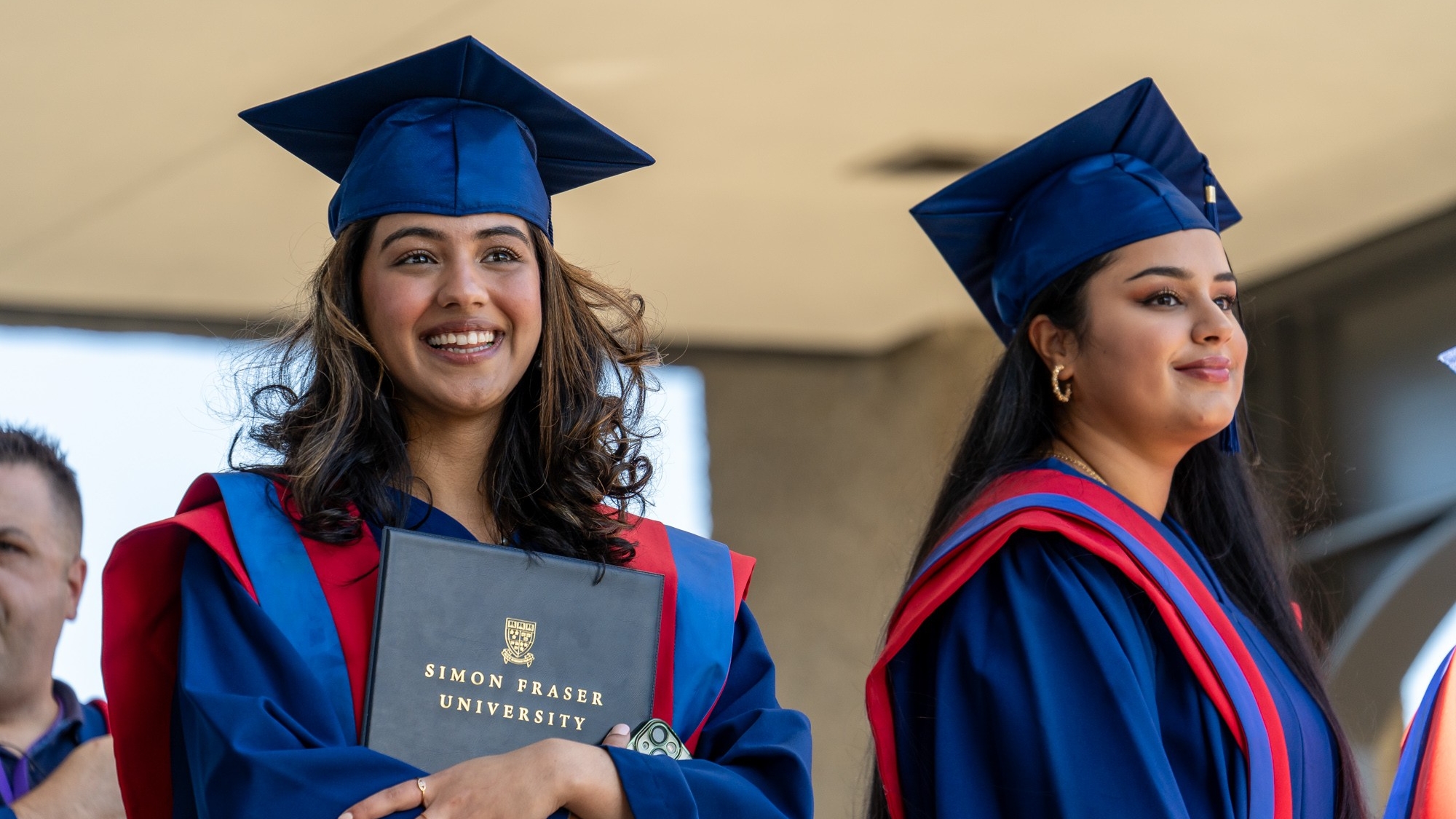 Two FASS graduates in the Convocation line up. The grad to the left is smiling and holding their parchment. The other grad to the right smiles off into the distance.