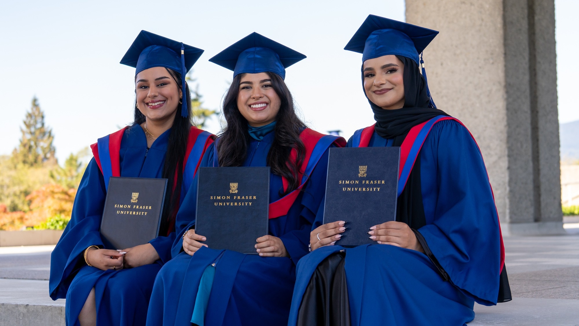 Three FASS graduates sitting on the steps of the AQ in their red and blue regalia. The graduates are showing their parchments and smiling at the camera.
