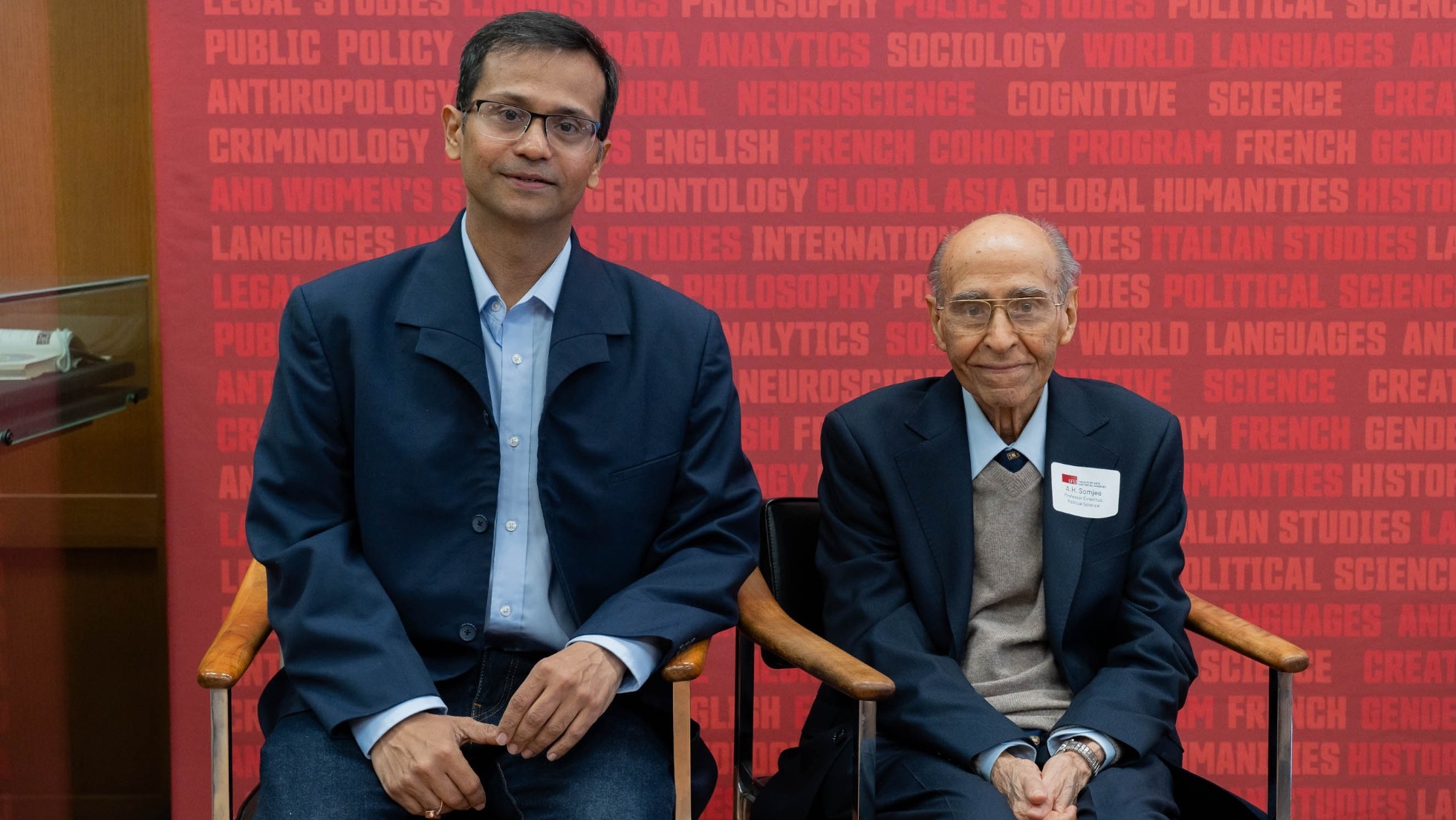 Shivaji Mukherjee and Dr. A.H. Somjee photographed sitting in front of the Faculty of Arts and Social Sciences banner at the Professorship launch event. Shivaji, pictured on the left, is has black hair and wears black glasses. He is wearing a light blue shirt with a navy coat. Dr. A.H. Somjee pictured on the right, smiles at the camera. He is wearing a brown sweater, a navy coat, and glasses.