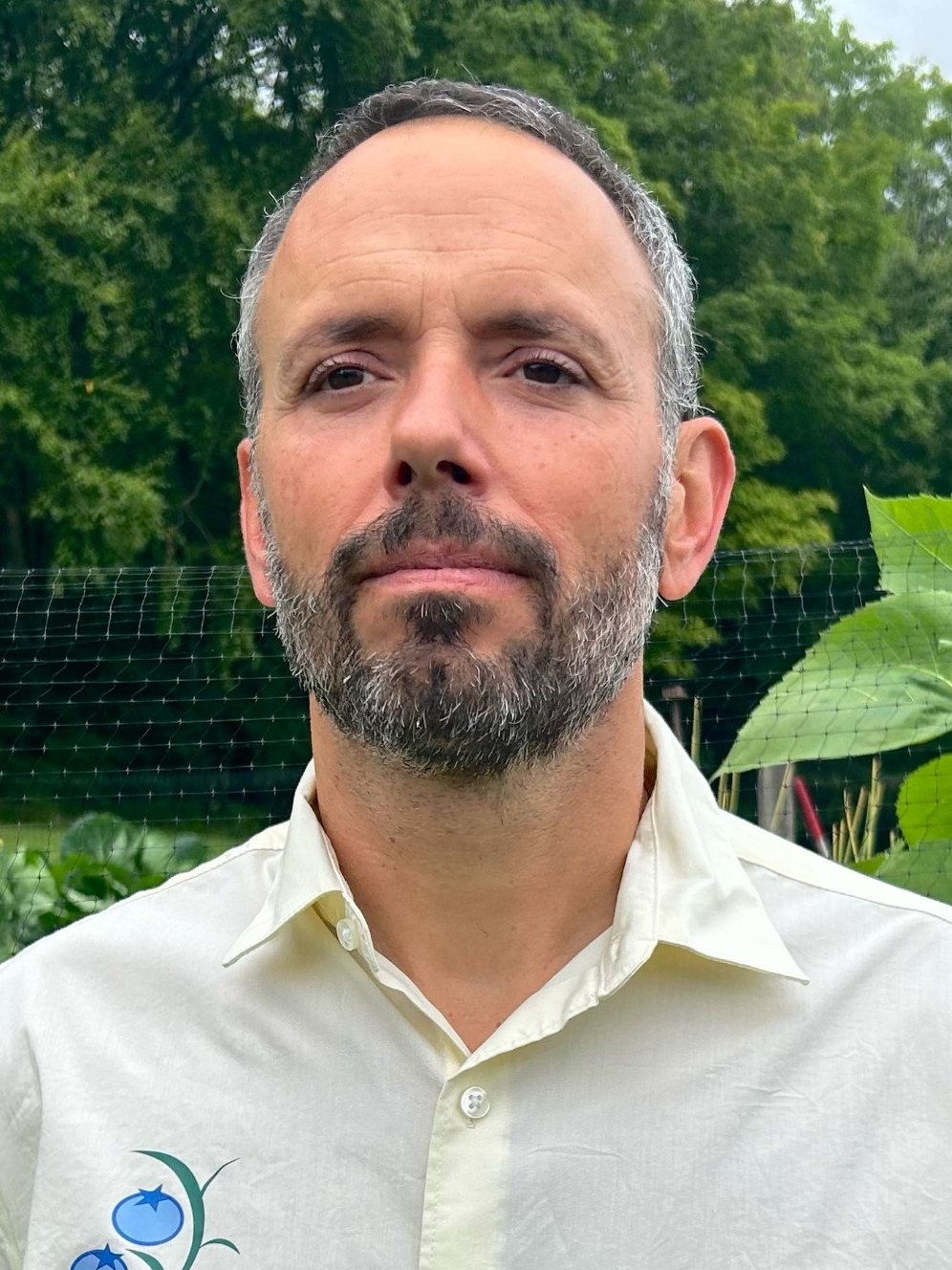 Nicholas is wearing a white button down shirt and is photographed outside in front of various greenery. Nicholas has short grey hair and dark grey facial hair.