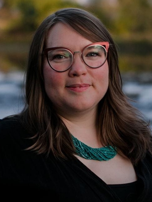 Zoe is photographed in front of a river. She has brown hair and is wearing red framed glasses with a large turquoise necklace.