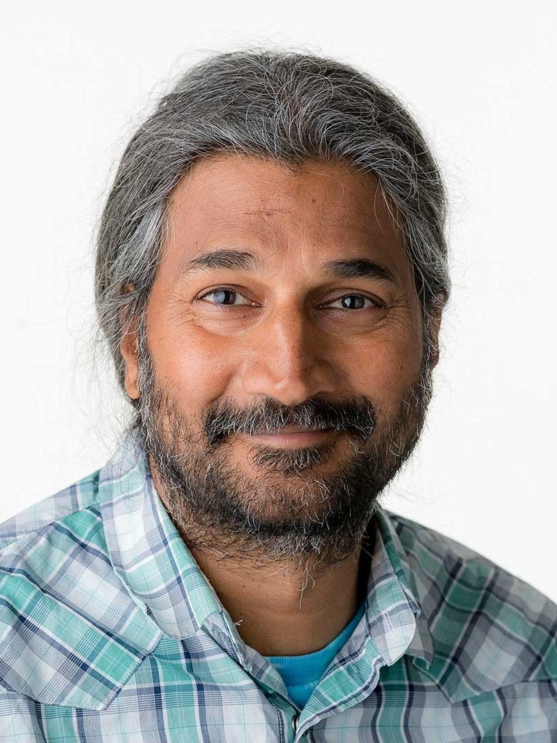 Krishna is wearing a blue plaid button down. He is photographed in front of a white background. He has dark grey hair and is smiling.