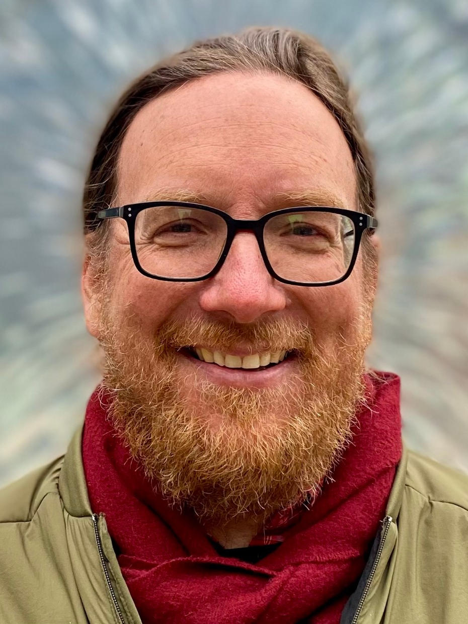 Michael is smiling at the camera with a red scarf around his neck. He wears black framed glasses, has light brown hair, and facial hair.