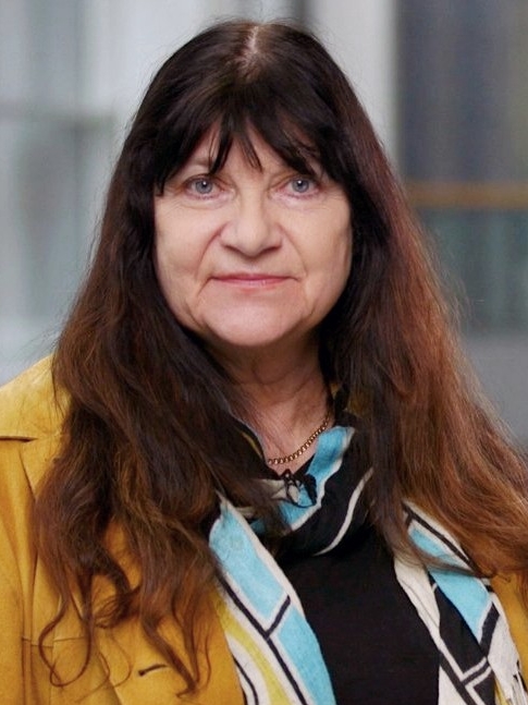 Portrait of Marianne wearing a bright yellow jacket smiling directly into the camera. She has long brown hair with bangs and blue eyes.