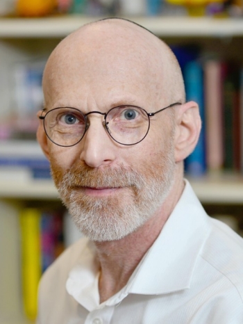 Terry is photographed in front of a library of books blurred in the background. He is a white man who has a short gray beard and is wearing black thin wire framed glasses and a white collared shirt. Terry is softly smiling directly at the camera.