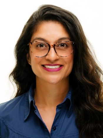 Roselene is a South Asian woman who is seen with wavy brown hair and wears large tortoise shell glasses. She is wearing a blue collared shirt and pink lipstick. Roselene is looking directly into the camera and smiling.