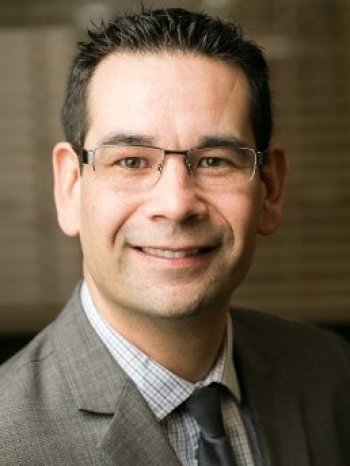 Mark wears thin framed glasses and has short brown hair. He is smiling directly into the camera for this portrait. He is wearing a white and blue plaid collared shirt with a black tie and a gray blazer.