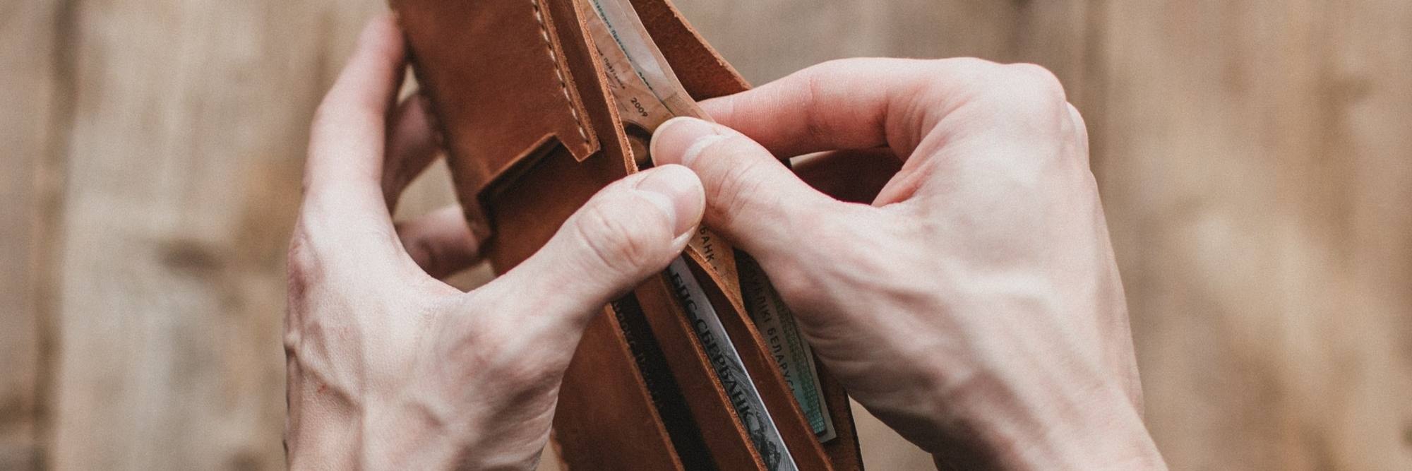 A person holding a brown leather bifold wallet in two hands holding onto a bank note.