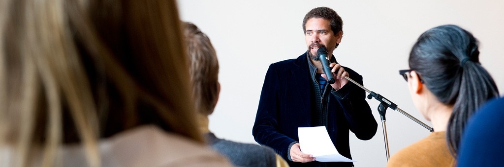 A man speaks into a microphone in front of a crowd who are blurred out in the foreground of the image. He has brown curly hair and brown facial hair. He is looking off into the distance while holding onto the microphone which rests on a microphone stand. To the left, white text overlaying red bars reads: LEARN THE ART OF PUBLIC SPEAKING.