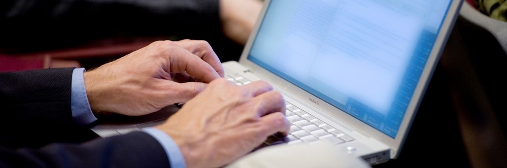 The hands of a person typing on a white MacBook Pro with black sleeves. On the left hand side in white text overlaying red bars reads: IMPROVE YOUR WRITING SKILLS.
