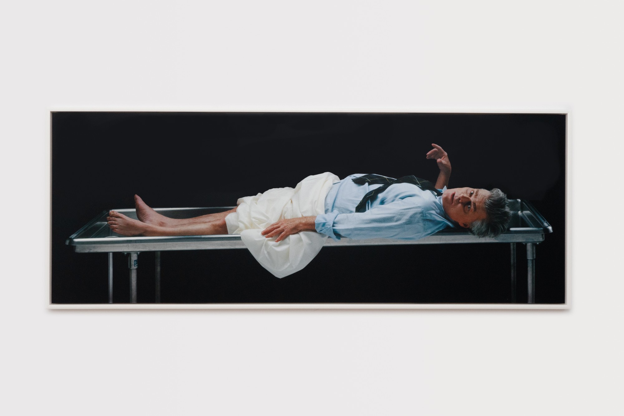 [Image Descriptions: A horizontal photograph depicts a middle-aged white man—the Canadian actor and filmmaker, Nicholas Campbell. He is lying face up upon a stainless steel mortuary table. Part of his upper body and head hangs off the edge of the rimmed table, and his gaze is oriented to the viewer. His legs and feet are bare from the knee down with a white sheet bundled over his waist and thighs. He wears a blue collared shirt and his black plaid tie is askance over his chest. His right hand points loosely upwards near his face: with eyes wide, brows raised and mouth slightly agape.]