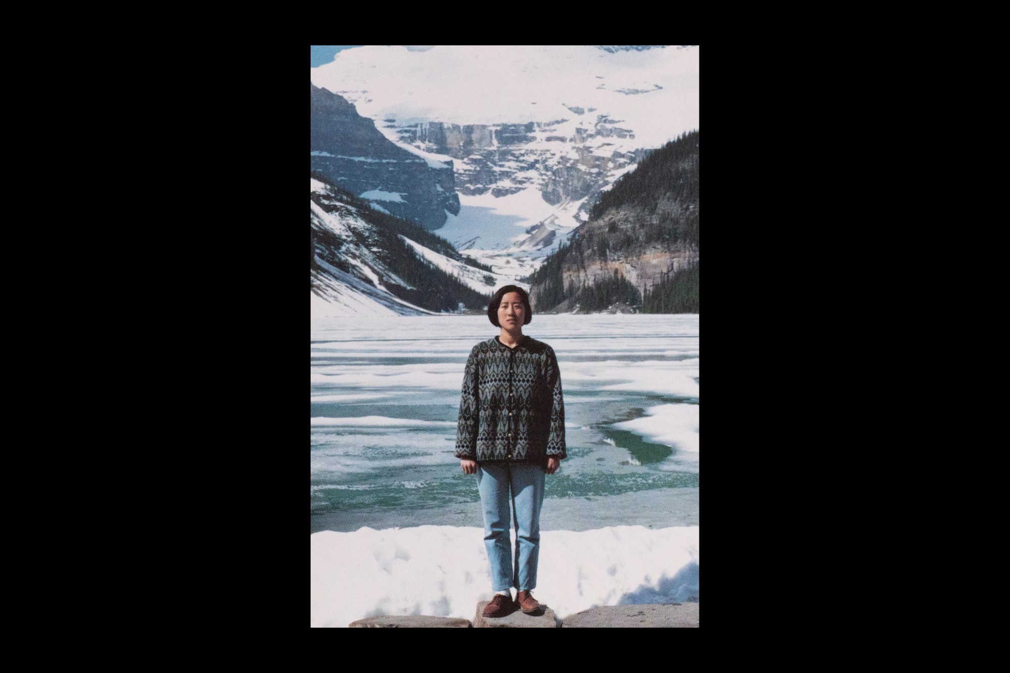 [Image Description: A postcard image shows the artist Jin-me Yoon standing before the frozen waters of Lake Louise and snow-covered mountains. She is wearing a dark patterned cardigan sweater, light blue jeans, and brown leather loafers. She stands erect on top of a stone, gazing directly at the camera with her hands forming loose fists by her side.]