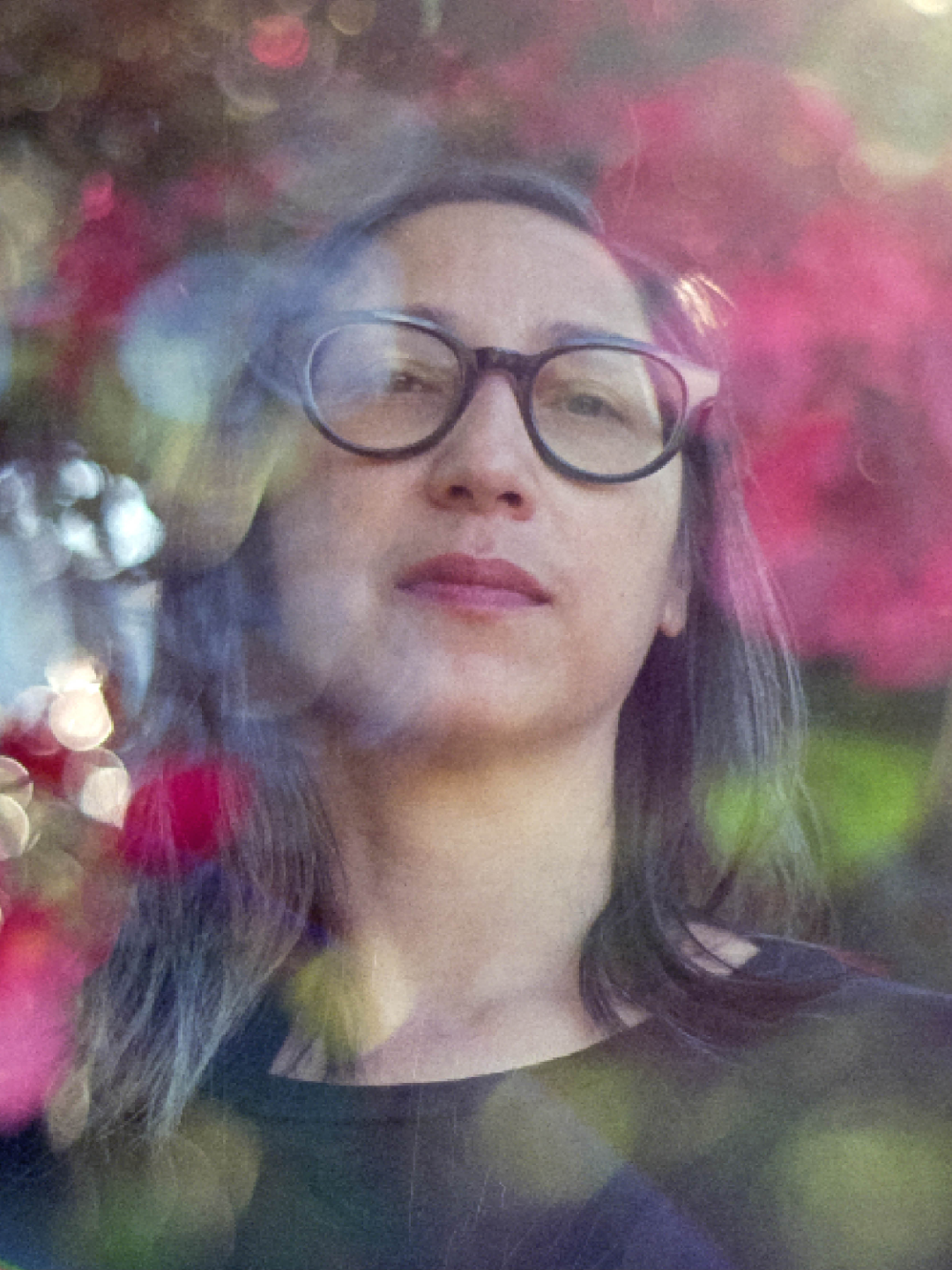 Mercedes Eng, a Chinese femme, wearing black plastic framed glasses and a black shirt is set amongst blurry and impressionistic flowering trees.