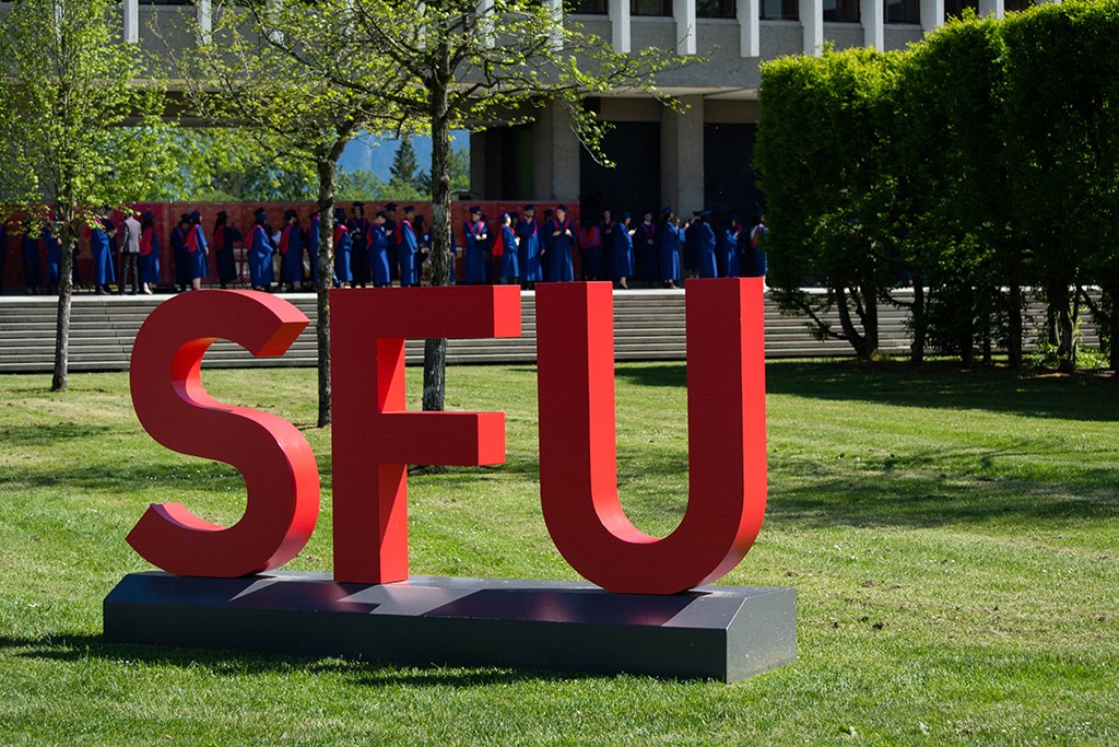 The SFU sign sits on the lawn of the AQ, with graduates in regalia lined up in the background. 