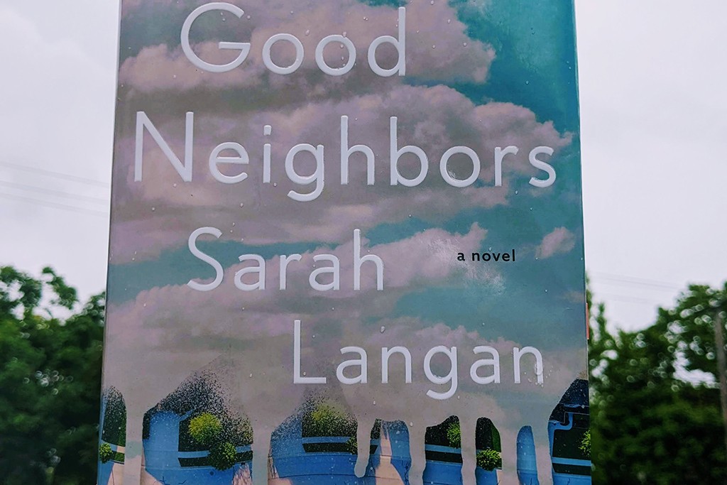 A hand holding up Good Neighbors by Sarah Langan, behind it is a cloudy sky and trees