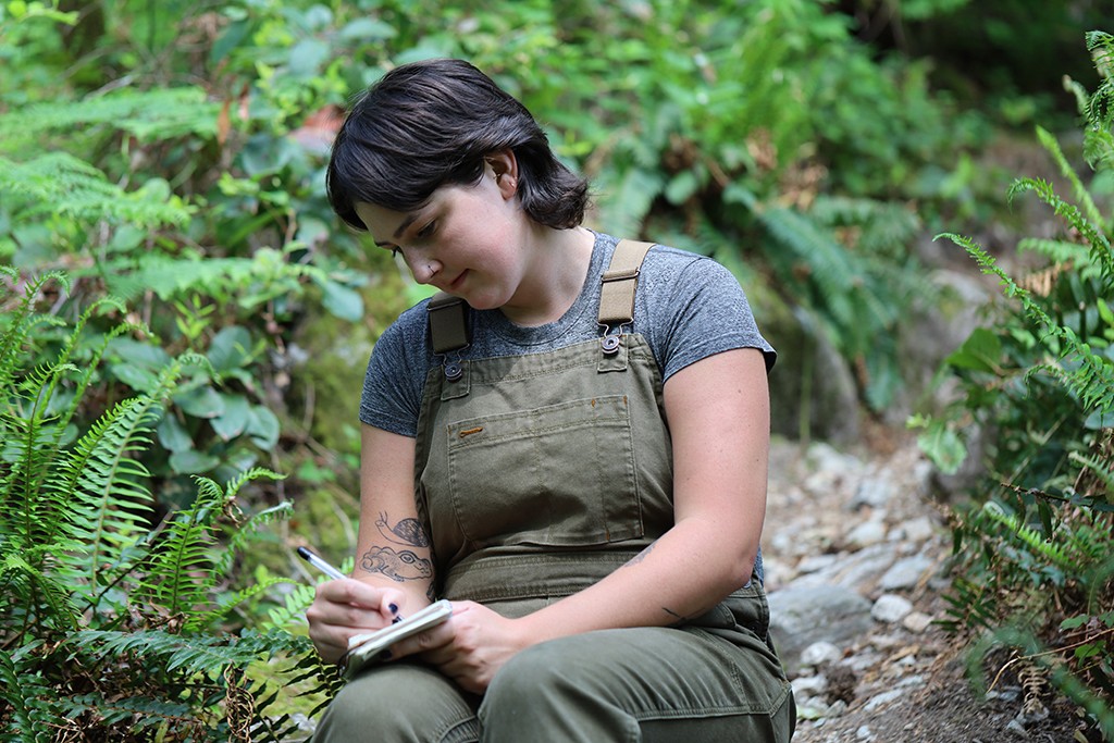 A woman with short brown hair sits in the forest, writing on a pad of paper