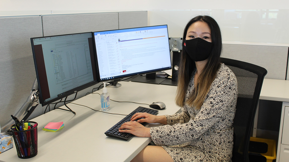 Joelle Mariano sits in a cubicle with two computer monitors in front of her. She has paused midway through typing and is looking at the camera, wearing an SFU-branded mask.