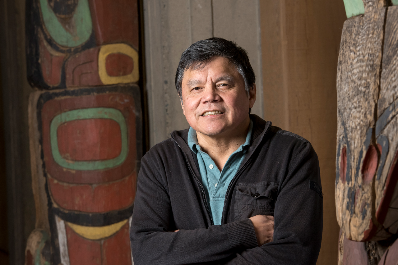 Gary George smiles at the camera with his arms crossed over his chest. He wears a blue-collared shirt with a black jacket over it. A wood wall and part of a totem pole are visible in the background.