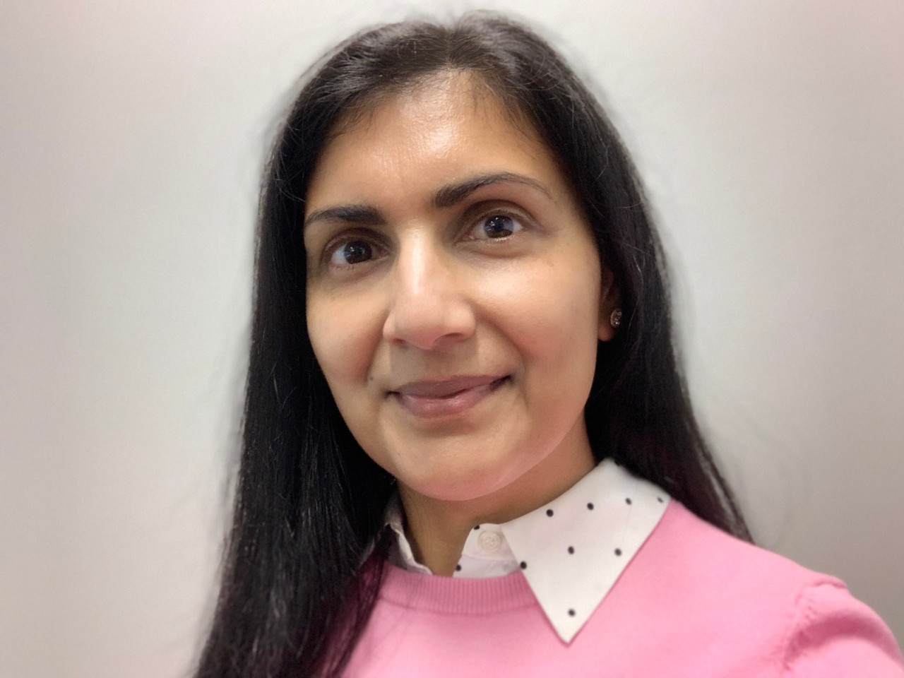 Suman Jiwani smiles at the camera against a grey backdrop. She is wearing a pink shirt with a white-and-black polka-dot collar.