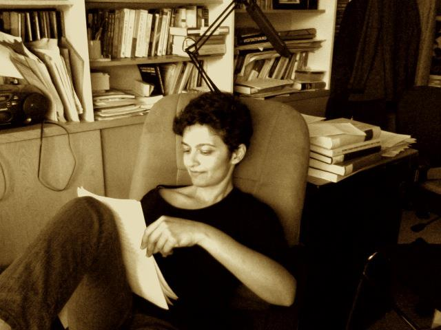 Nilima Nigam in her office. She is leaning back in her chair and reading the papers propped up on her leg. There are cabinets, bookshelves, and a coat in the background.