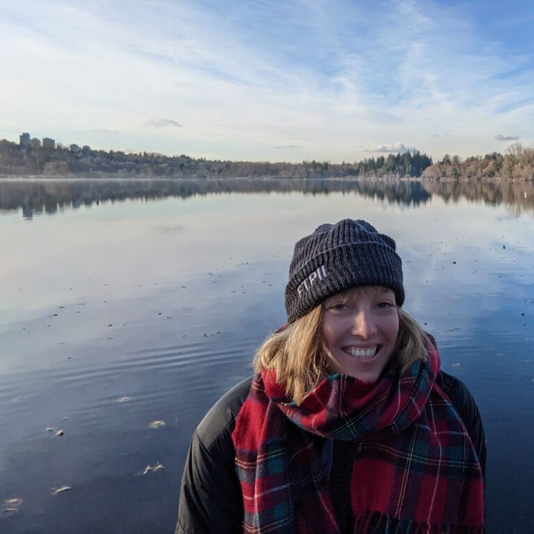 Nicola Mulberry smiling in front of a blue body of water. There are trees, white clouds, and a blue sky on the horizon that are reflected on the water.