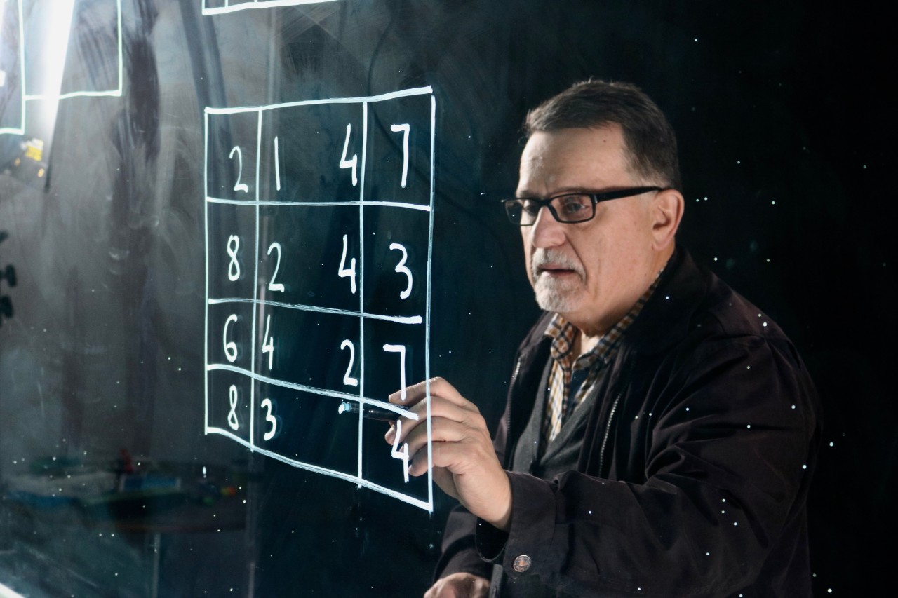 Veselin Jungic solving a puzzle with a writing tool. The puzzle is in white ink on a transparent surface. It consists of a square with intersecting lines and numbers between the lines. 