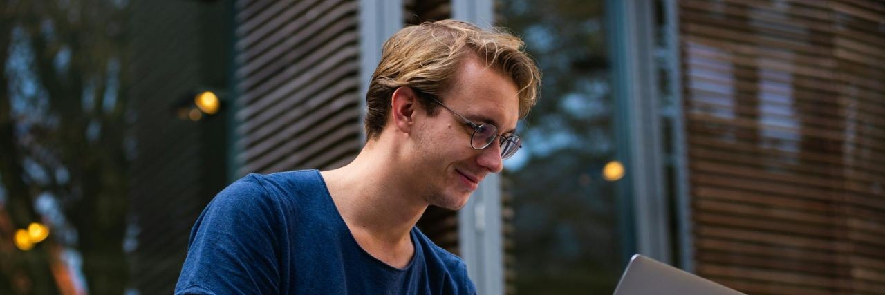 A person wearing glasses and a blue shirt sits outside using a laptop, with a building and trees in the background.