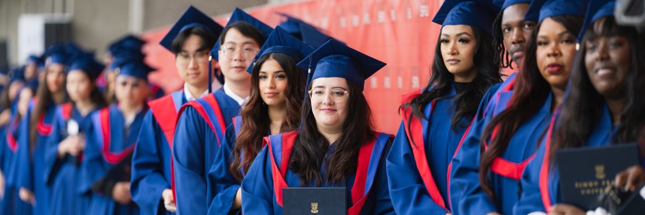 A group of graduates in blue caps and gowns stand in a line holding Simon Fraser University degrees.