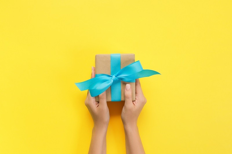 Woman hands give wrapped valentine or other holiday handmade present in paper with blue ribbon. Present box, decoration of gift on yellow table, top view with copy space