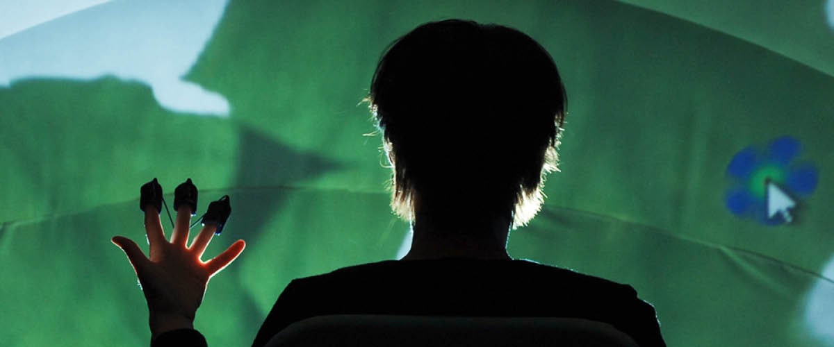 Photo of a silhouetted person in front of a screen wearing virtual reality motion-tracking devices on their fingertips.