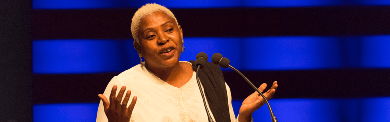 Dr. Afua Cooper speaks at a podium in front of a blue theatre curtain