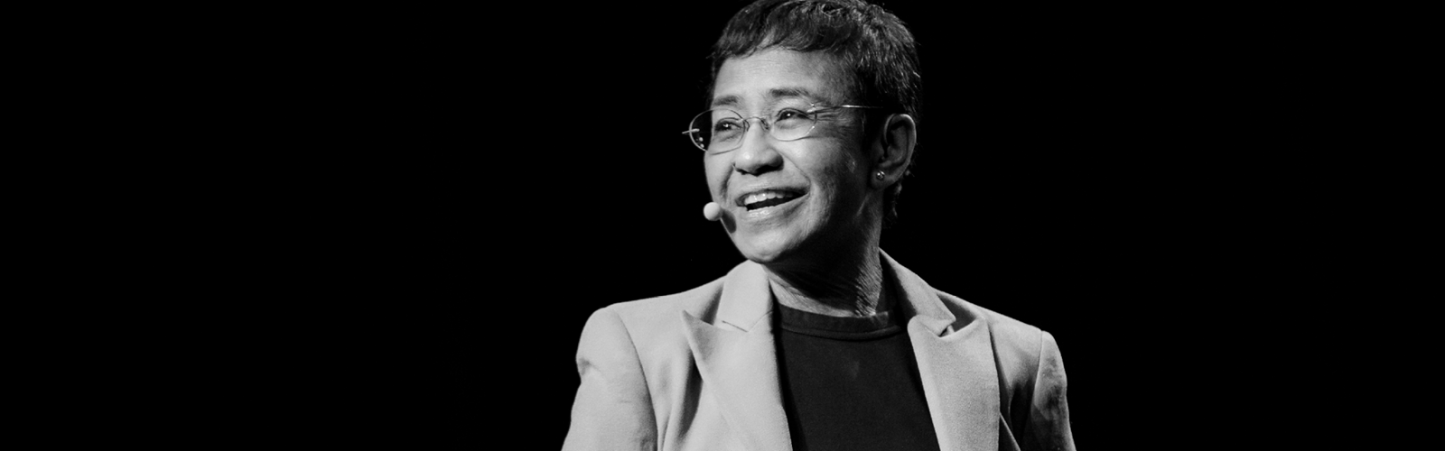 Maria Ressa smiling on a dark background wearing a headset microphone during a past speaking event.