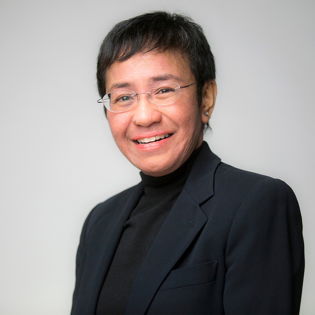 Maria Ressa bio photo shows Ressa smiling on a light grey background while looking toward the camera