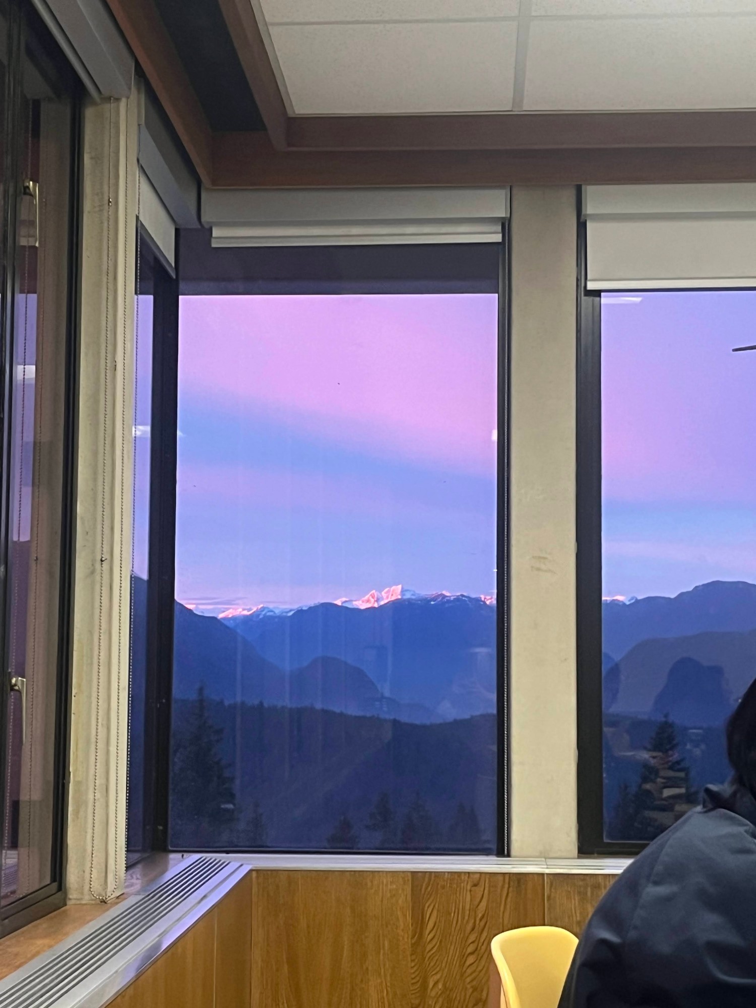 A window on SFU Burnaby campus overlooking a purple and blue evening sky, with the mountains in the far background and the back of a student seated in the foreground.