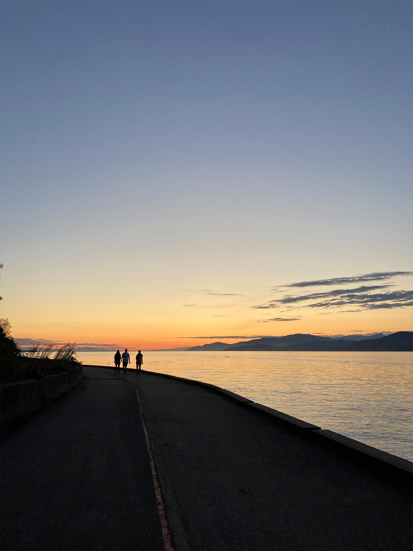 The seawall in Stanley Park next to a sun setting into the ocean and mountains beyond.