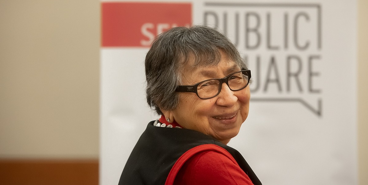A photo of Elder Margaret George of the Skawahlook First Nation, dressed in black and red, looking back at the camera and smiling in front of a sign with the SFU Public Square logo on it.