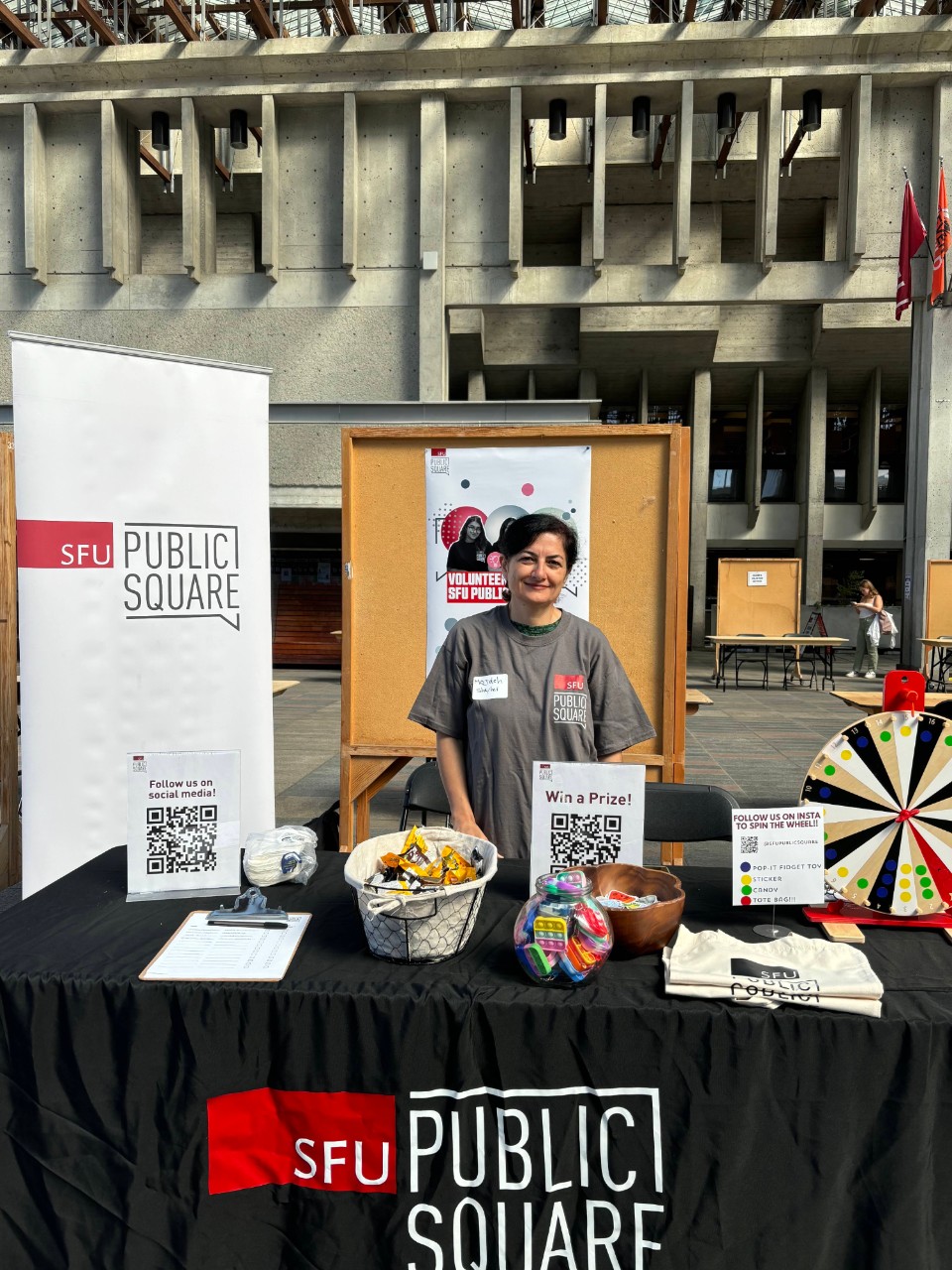 An image of Mozdeh Eftekhari volunteering at the SFU Ready Fair Burnaby. She is standing in front of a table with a SFU Public Square's logo on a poster behind her. On the table there are toys, stickers, and a spinning prize wheel.