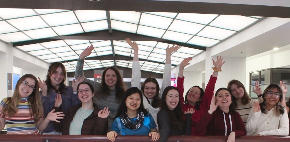A group of eleven of our 2024-2025 graduate students standing together waving from the second floor inside the Downtown Vancouver campus of SFU.  