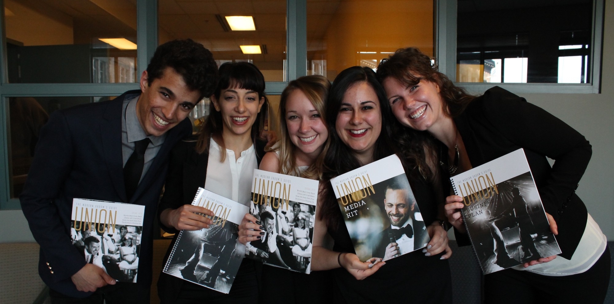 Five Master of Publishing students standing closely together, smiling and holding their magazine projects