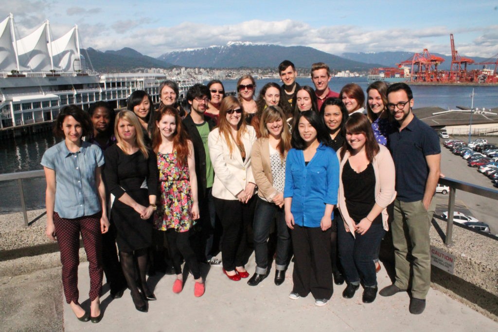 A group of approximately 20 graduate students in the Master of Publishing program standing together. They are in downtown Vancouver, the waterfront and mountains are visible in the background.