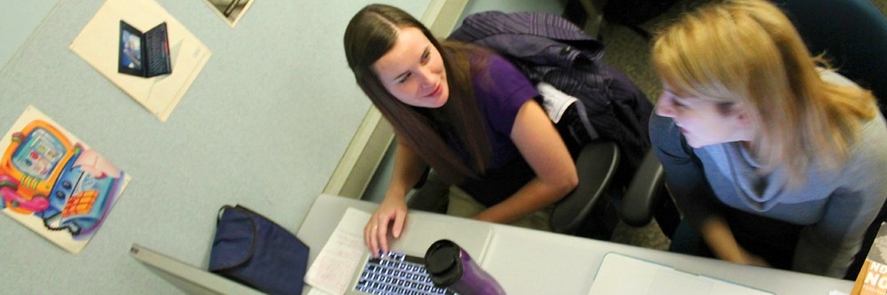 Two students sitting at a desk, working at their laptops and chatting
