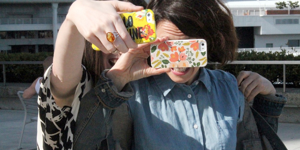 Two women stand closely together to take a selfie, holding up their smartphones up, blocking their faces.