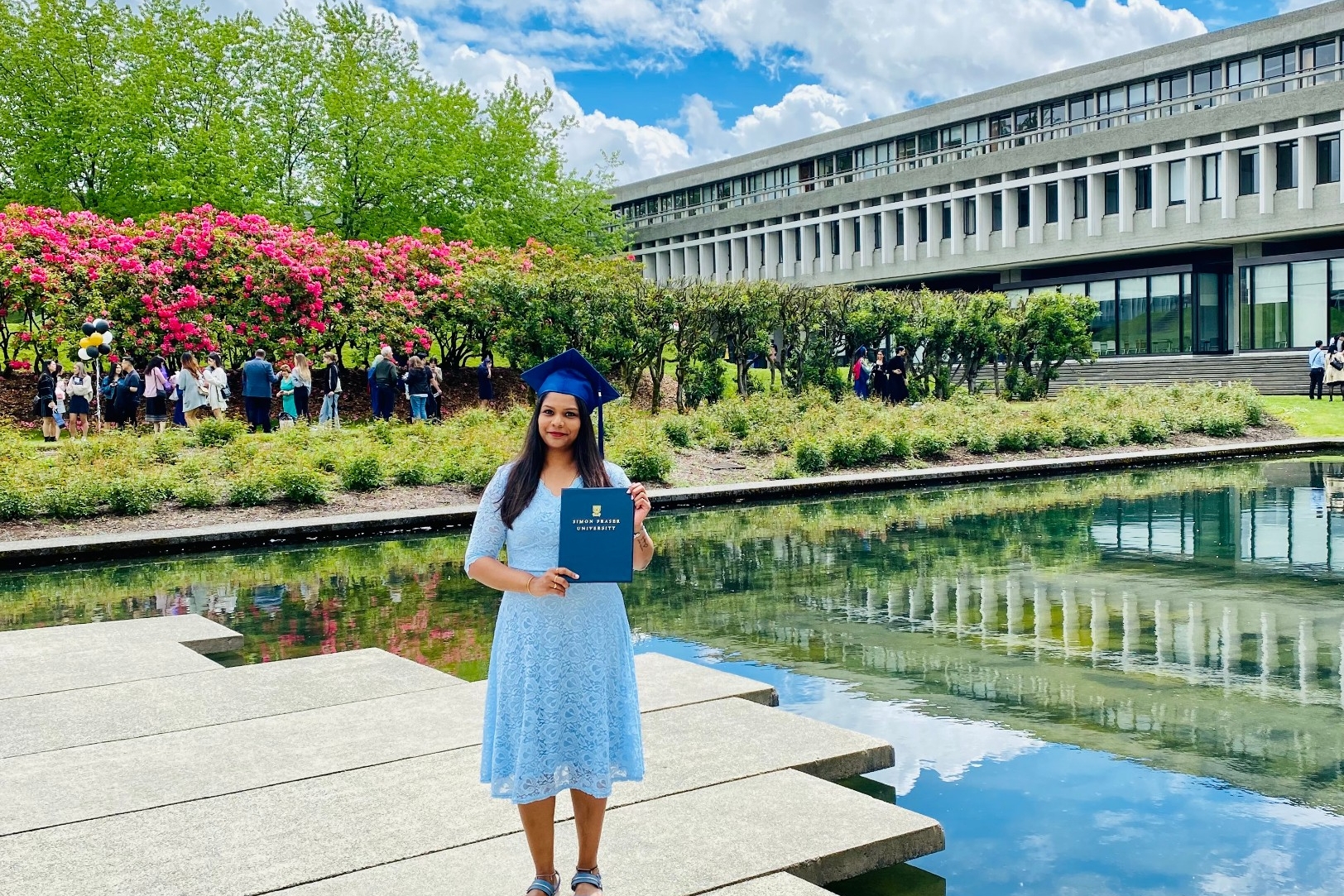 MPub alumnus Christina Paul with her Masters degree in front of the AQ pond at SFU's Burnaby campus. She is donning a bright floral dress and her graduation cap.
