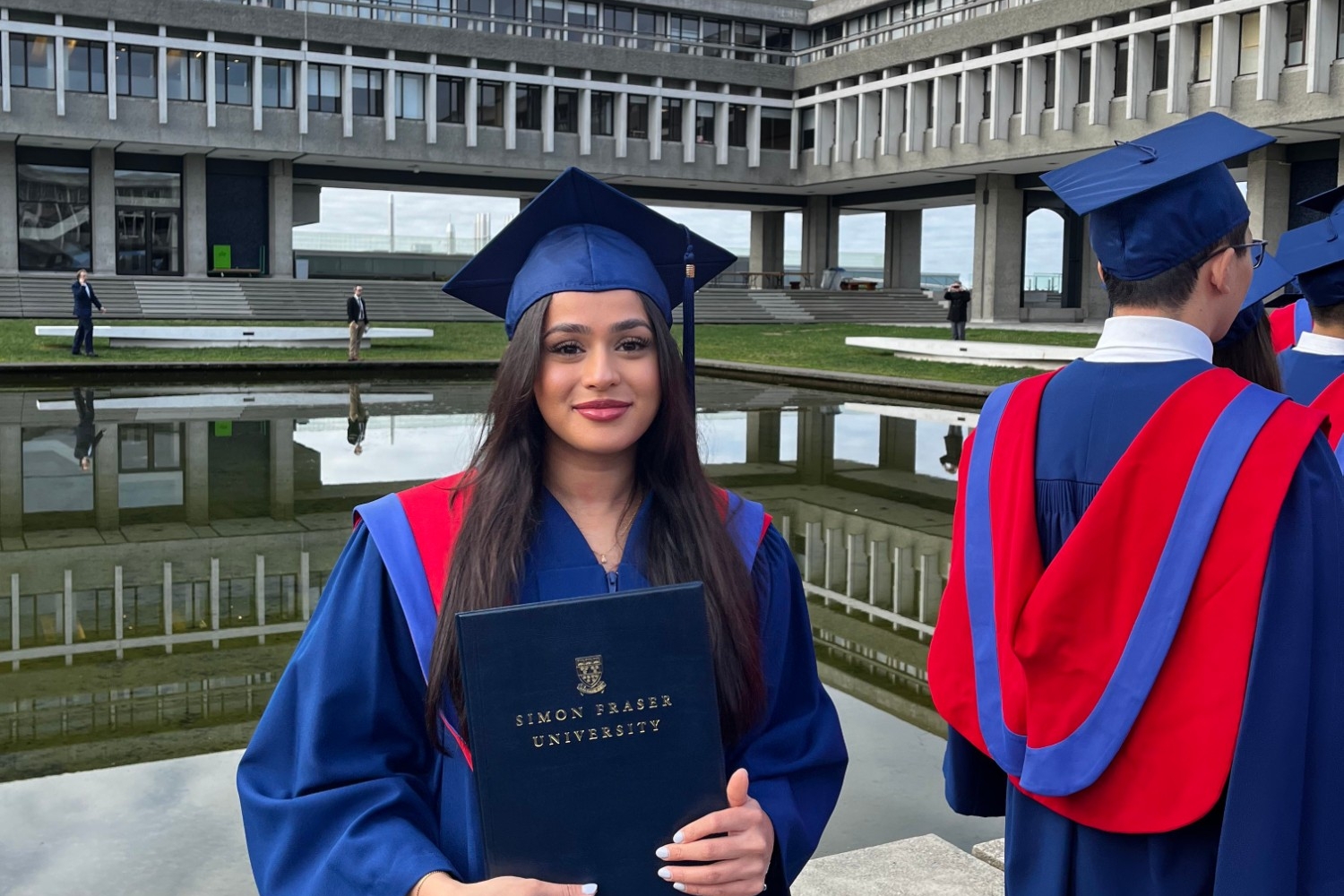 Marihah Hussainaly Farook in full regalia at her convocation holding her parchment as she poses in front of SFU's reflection pond.