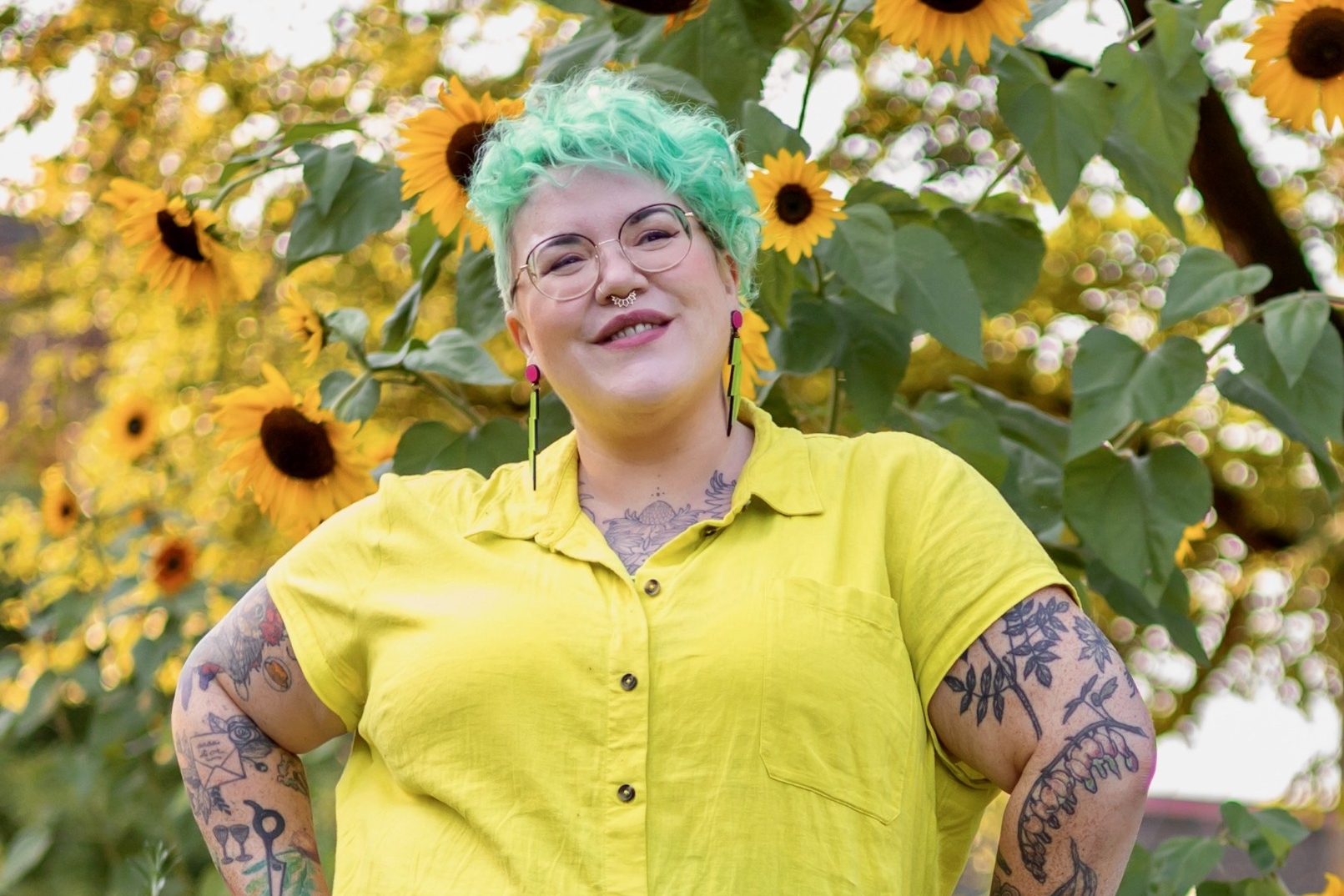 Hannah in a bright pear coloured shirt dress gleefully posing in front of a sunflower tree with both her hands on her hips. 